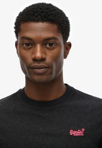 Zwarte katoenen t-shirt met een ronde halslijn, voorzien van een klein, roze geborduurd "SuperDry" logo op de linkerkant van de borst. Soepele textuur.