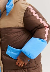 Hnedá a béžová zateplená bunda s jasne modrými manžetami a kapsičkou, ružový vlnitý pruh na rukáve, nosená osobou s rukou pri pocket.