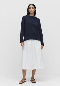 Marinblå stickad tröja med ribbat nederkant och långa ärmar, kombinerad med en svängig vit midiklänning och bruna platta sandaler. Minimalistisk design.