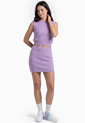 Vrouw draagt een lavendelkleurige mouwloze gebreide top en bijpassende rok met witte sokken en witte sneakers met pasteltinten, staand en glimlachend.