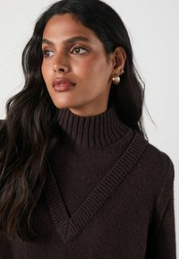 Brown turtleneck sweater with a ribbed texture and V-shaped neckline. Model has wavy hair and wears gold hoop earrings.