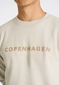 Sudadera color beige claro con las palabras "COPENHAGEN" y "LINDBERGH" impresas en colores contrastantes. Tejido suave con cuello redondo.