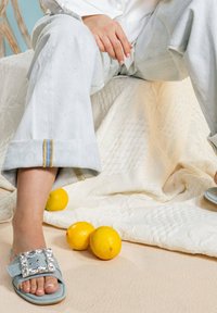 Lichtblauwe sandalen met een vierkante strass gesp, gedragen met lichtgrijze, losse broek met een gestikt patroon en gele accenten.
