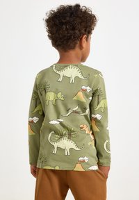 Maglietta a maniche lunghe verde con stampa di dinosauri di varie specie, alberi e vulcani. Il materiale sembra morbido e confortevole.