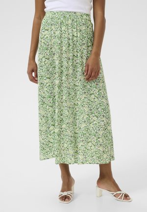 Femme portant une jupe midi fleurie verte et blanche, un haut blanc sans manches et des sandales blanches à talons avec des brides sur un fond uni.