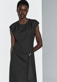 Schwarzes, ärmelloses Kleid mit strukturierten Schulterdetails, einem drapierten Vorderteil und einem silbernen Akzent an der Taille. Hergestellt aus einem strukturierten Stoff.