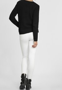 Svart texturerad långärmad skjorta kombinerad med vita åtsittande jeans och svarta ankelboots med nitar och spännen, sedd bakifrån.