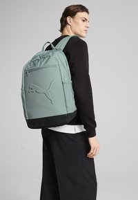 Roheline riidest seljakott, millel on silmatorkav reljeefne Puma logo, võrkkotid külgedel, reguleeritavad sangad ja lame must alumine paneel.