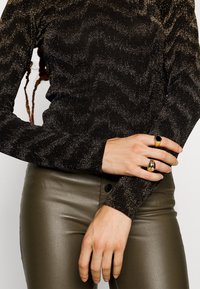 Schwarzes Langarmoberteil mit metallischem Glanz und Wellenmuster, kombiniert mit olivgrünen, figurbetonten Hosen. Zwei goldene Ringe an der Hand.
