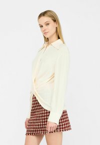Blusa color crema con un dettaglio annodato in vita, maniche lunghe e colletto classico, abbinata a una gonna a pied de poule rossa.