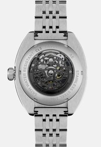 Montre en acier inoxydable avec un fond de boîtier transparent révélant des mouvements mécaniques noirs complexes et des accents colorés de bijoux. Étanche.