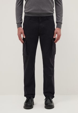 Uomo che indossa pantaloni cargo neri a gamba dritta con tasche a patta e scarpe nere con lacci, abbinati a un maglione grigio a maniche lunghe.