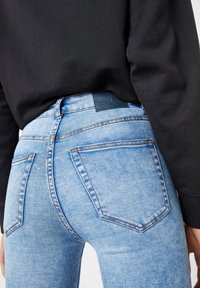 Jean skinny en denim bleu clair à taille haute, doté de cinq poches, d'une texture douce et de surpiqûres contrastantes. Étiquette de ceinture noire visible.