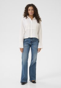 White button-up shirt with pleating detail, paired with light blue flared jeans. Shirt features long sleeves with wide cuffs and a fitted waist.