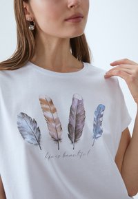 White short-sleeve t-shirt featuring watercolor feather prints in blue, brown, and gray, with the text "life is beautiful" beneath.