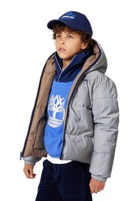 Gutt som har på seg en blå caps, blå sweatshirt med hvitt tre-logo, grå dunjakke og mørke bukser, ser litt oppover og mot høyre.