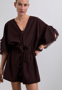 Mujer vestida con un vestido suelto de color marrón oscuro con cuello en V, cintas con borlas, mangas cortas deshilachadas y una pulsera negra en su muñeca derecha.