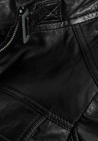 Close-up van een zwart leren jack met een textuur, stikdetails, metalen gesp en rits.