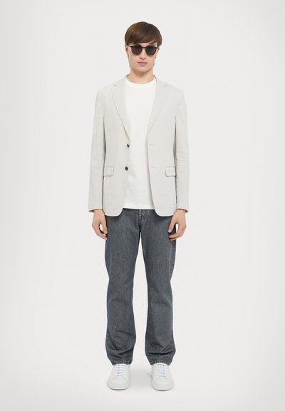 Hellgrauer, taillierter Blazer, weißes T-Shirt, dunkelgraue Jeans und weiße Sneaker. Runde schwarze Sonnenbrille; minimalistische Stil mit klaren Linien.