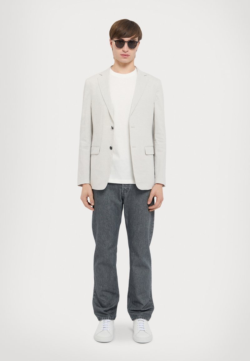 Blazer grigio chiaro su misura, t-shirt bianca, jeans grigio scuro e sneakers bianche. Occhiali da sole neri rotondi; stile minimalista con linee pulite.