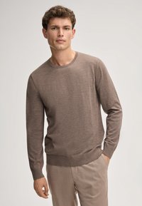 Brauner Strickpullover, Rundhalsausschnitt, lange Ärmel, gerippte Bündchen und Saum, weiche Textur, getragen mit hellbeigen Hosen. Keine sichtbaren Muster oder Logos.