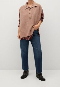 Virkat rosa ärmlös topp med krage och tre träknappar, lager-på-lager över en röd och vit randig skjorta. Blå smala jeans och svarta klackstövlar.
