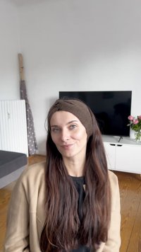 Καφέ πλεκτή κορδέλα φορεμένη από άτομο με μακριά σκούρα μαλλιά, σε ένα δωμάτιο με μια σκοτεινή τηλεόραση και μια ανθοδέσμη με ροζ λουλούδια.