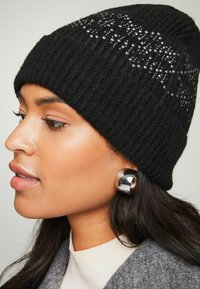 Gorro de punto negro con canalé y detalles de strass. Presenta una textura suave y un ajuste ceñido. Combinado con una blusa de color claro y pendientes de plata.