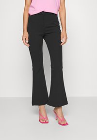 Kvinna i svarta utsvängda byxor, rosa remsandaler med hög klack och en ljusrosa topp, står mot en enkel ljus bakgrund.