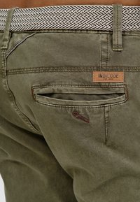 Oliivinvihreät housut, joissa on nahkainen merkkilaatta, kudottu kuvioitu vyö ja takatasku, jossa on kirjailtu koriste.