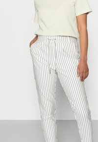 Valkoiset raidalliset housut, joissa on mustat pystysuuntaiset viivat, joustovyötärö, etutaskut ja rento leikkaus. Yhdistetty vaaleanvihreään yläosaan.