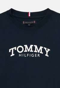T-shirt de gola redonda em azul marinho com o texto "TOMMY HILFIGER" em branco centrado no peito e um pequeno rótulo vermelho, branco e azul dentro do colarinho.