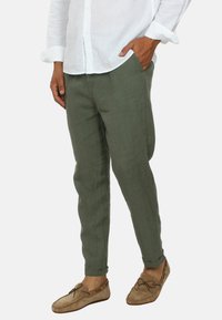 Pantaloni in lino verde oliva con una vestibilità affusolata, tasche laterali e orli risvoltati, abbinati a scarpe beige slip-on e una camicia bianca con bottoni.