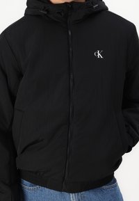 Czarna kurtka z kapturem zapinana na zamek, z elastycznym pasem, bocznymi kieszeniami i białym logo "cK" na lewej piersi, noszona na niebieskich dżinsach.