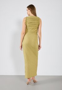 Robe maxi verte à fronces avec un tissu texturé et sans manches, présentant un dos froncé et une fente sur le côté. Sandales à talons blocs transparentes.