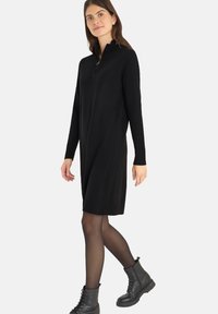 Abito nero in maglia con colletto a metà zip, tessuto a costine sulle maniche e una vestibilità comoda, abbinato a stivali combat neri e collant trasparenti.