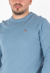 Blauwe langwerpige shirt met lange mouwen, gemaakt van ribbelstof, met een ronde hals. Heeft een klein geborduurd vlaggetje op de borst.