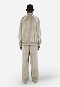 Bársonyos tréningruha világos bézs színben, fehér oldalsávokkal. Magas gallérral és bordás mandzsettákkal rendelkezik. Laza fazon, széles lábakkal.