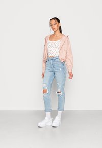 Jacket bomber rosa, top branco com padrão de cerejas, jeans rasgados de um azul claro e tênis brancos chunky. Modelo posando contra um fundo liso.