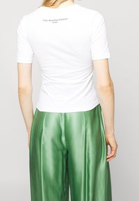 Camiseta blanca ajustada de manga corta, con un pequeño logo impreso en la parte posterior. Combinada con pantalones anchos verdes brillantes.