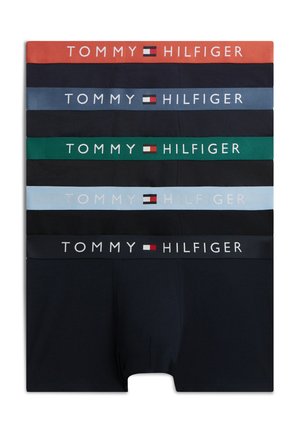 Joven de pie descalzo vistiendo bóxers azul marino de Tommy Hilfiger sobre un fondo blanco liso.