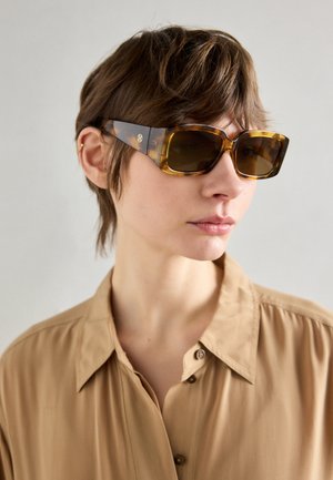 Óculos de sol em padrão tartaruga castanho com lentes retangulares grandes e lados grossos, apresentando uma marca discreta numa das hastes. Acabamento suave e brilhante.