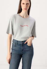 Grå t-skjorte med korte ermer med rød brodert tekst "Be Savage Not Average." Kombinert med mørkeblå jeans.