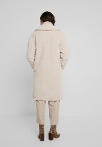 Lång beige kappa med en mjuk, texturerad yta och en fluffig krage. Bärs över krämfärgade byxor och mönstrade ankelskor med klack.