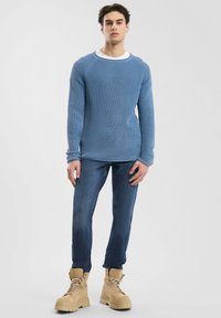 Blauer gestrickter Pullover mit Rundhalsausschnitt, kombiniert mit dunklen Jeans und beigen Schnürstiefeln. Die Texturen sind ausgeprägt und übereinander geschichtet.