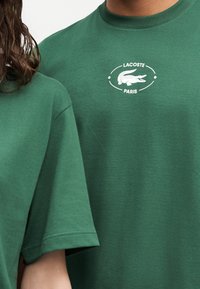 Δύο άτομα φορούν σκούρα πράσινα T-shirt Lacoste, με ένα T-shirt να φέρει το λευκό λογότυπο Lacoste Paris με έμβλημα κροκοδείλου.