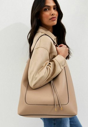 Mujer con chaqueta beige que sostiene un gran bolso tote de cuero beige con correa negra y flecos sobre el hombro.