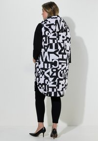 Svart och vit vadderad väst med stora grafiska bokstäver, huva, sidoslitsar och en längre baksida, som bärs över en svart topp och leggings.