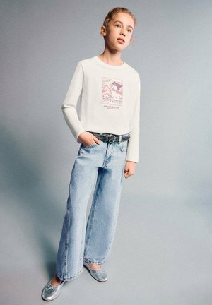 Camicia bianca a maniche lunghe con stampa grafica rosa, jeans denim azzurri a gamba larga e scarpe piatte argento. Cintura nera semplice con fibbia decorativa.