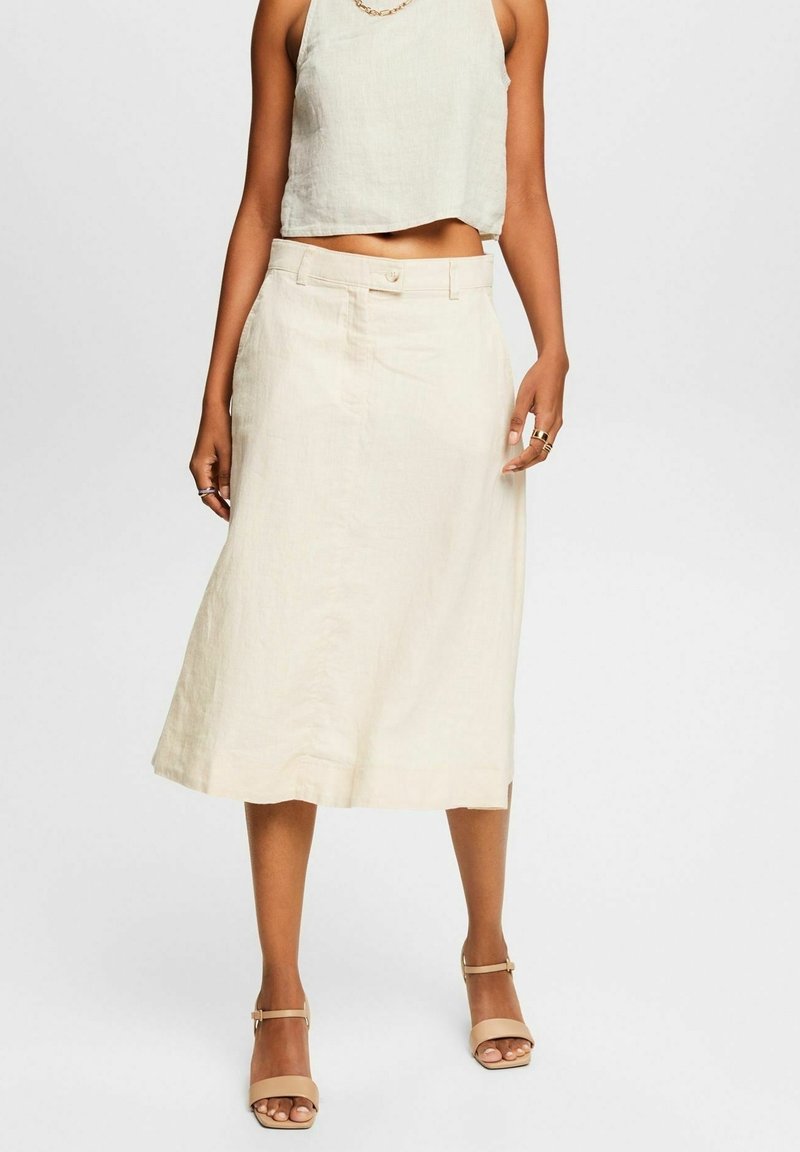 Femme portant un top court écru sans manches, une jupe midi taille haute, un collier chaîne en or, des bagues et des talons ouverts beige.